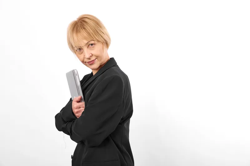 Elderly woman, white background, studio, holding hair brush, smiling softly, black jacket, short blonde hair, side pose, minimal backdrop, quiet mood