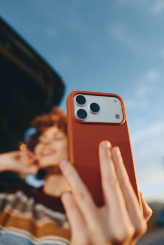 Woman lifestyle smartphone hands selfie outdoors, orange phone case close up, blurred smiling background shows couple, casual summer moment, capturing memories with technology, shallow depth of field