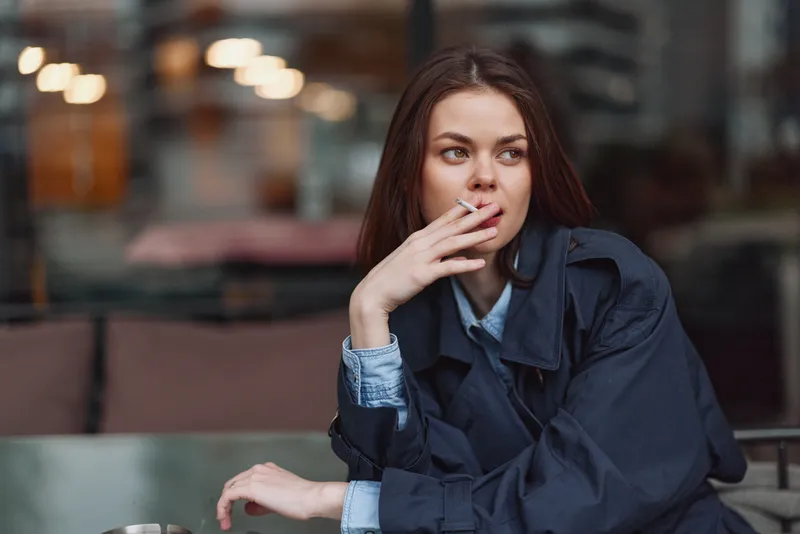 Young woman in a denim shirt and dark coat sits at an outdoor cafe, cigarette in hand, thoughtful gaze, moody natural light, casual urban moment.