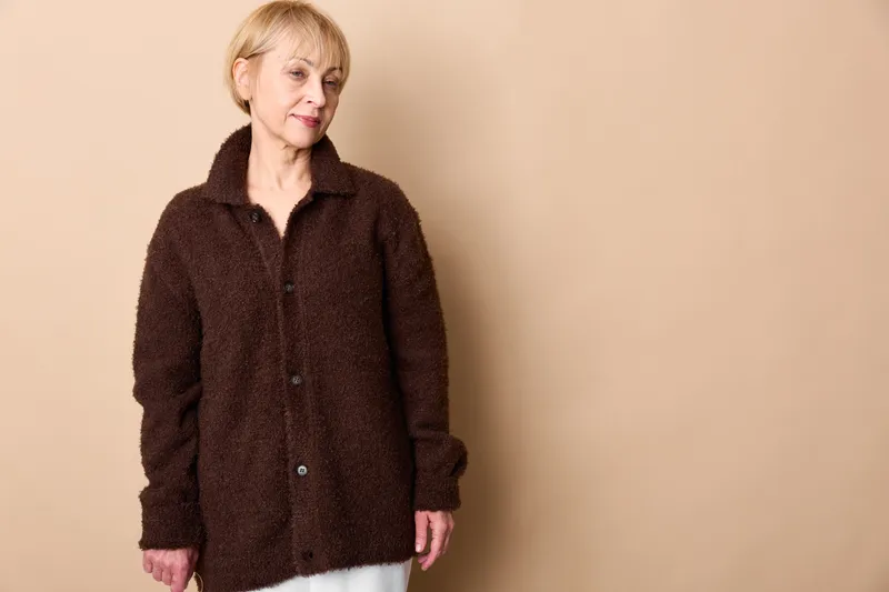 Elderly woman standing in dark coat against beige wall, neutral expression, studio portrait, soft light, calm mood, minimal background, copy space, indoors, mature demeanor, pose