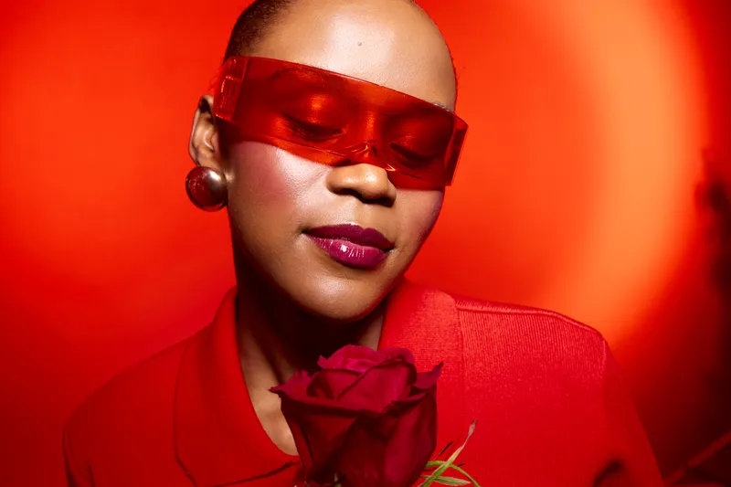 Close-up of a dark skinned African woman with shaved head wearing red sunglasses and red outfit holding a red rose. Concept of beauty, fashion, and self-expression on red background.