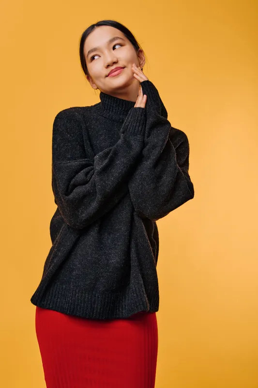 Young woman in black turtleneck sweater and red skirt posing thoughtfully against orange background with hands near face and slight smile for fashion portrait.