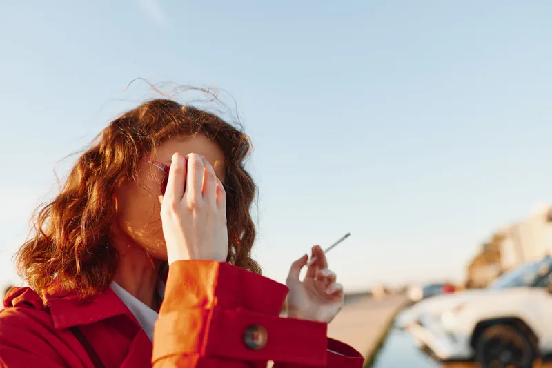 Young woman with curly hair wearing a red coat adjusts her sunglasses outdoors on a sunny day with a blurred urban background.