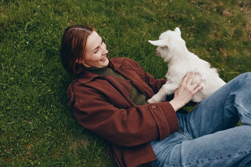 Relaxed woman lying on grass wearing a brown jacket, holding a white goat kid, smiling while they share a gentle moment outdoors in a sunny meadow, natural lifestyle scene