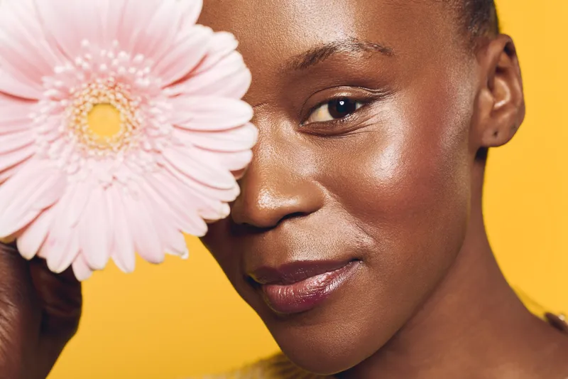 Close-up of a smiling African woman with dark skin holding a pink flower near her face. Yellow background and soft lighting create a warm, natural beauty and skincare concept.