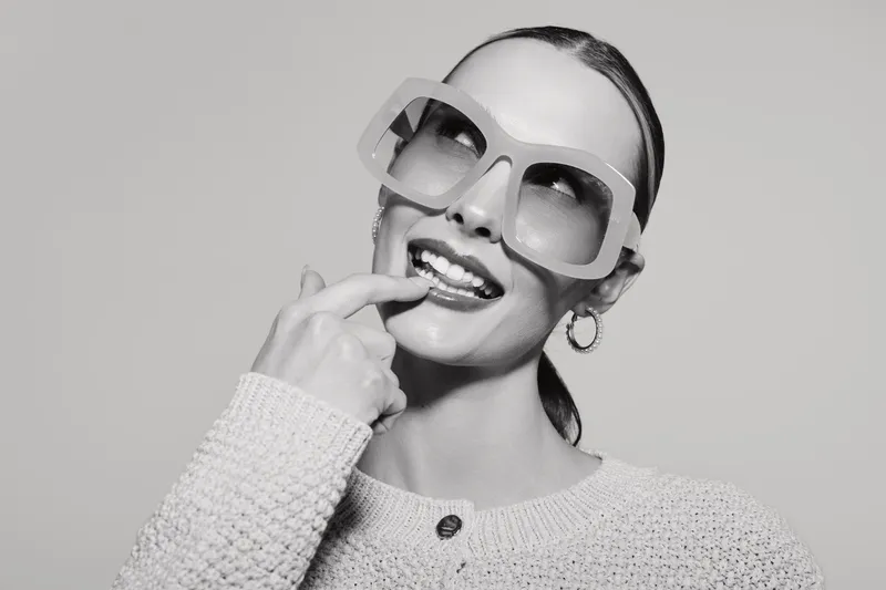 Happy young caucasian woman with light skin wearing oversized sunglasses and knit sweater smiles playfully, touching her lips. Studio portrait in black and white with fun and fashion concept.