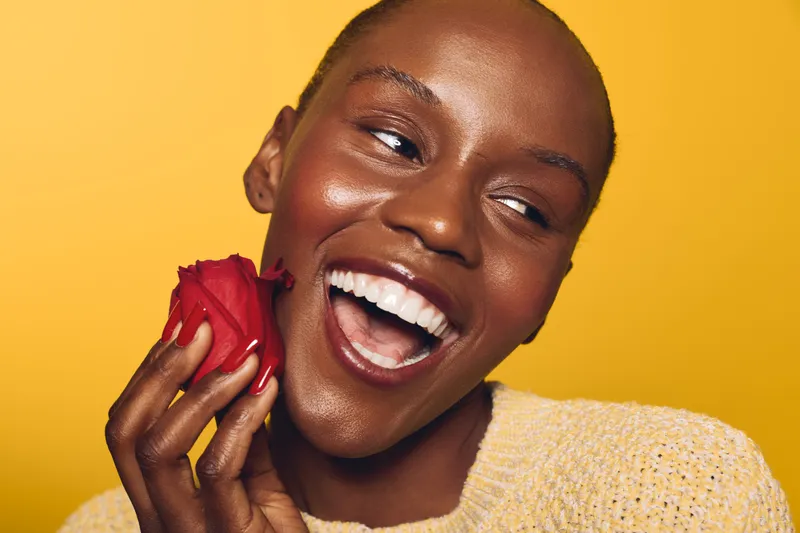 Happy African American woman with dark skin smiling and holding a red rose near her face on yellow background. Concept for beauty, joy, and natural skincare with floral element.