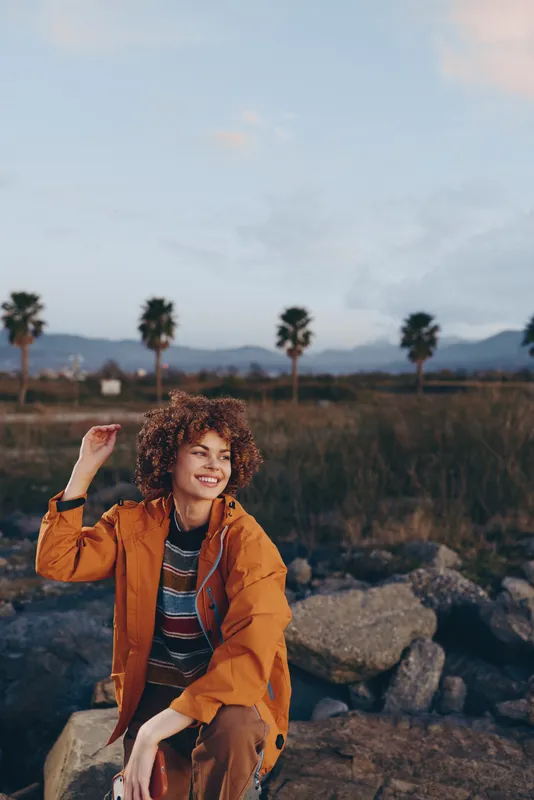 Woman with curly hair, outdoors in orange jacket, smiling portrait in rocky nature, palms and distant mountains in background, lifestyle travel moment, soft sunset light, calm mood