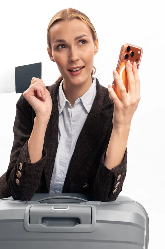 Young woman with blonde hair holds smartphone and suitcase, smiling and gesturing while planning travel. Concept of tourism, vacation, technology, and communication. Bright studio lighting.