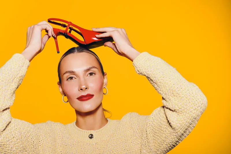 Young woman in beige sweater holds red high heel shoe above head on bright yellow background. Concept for fashion, style, footwear, and beauty with vibrant colors and confident expression.