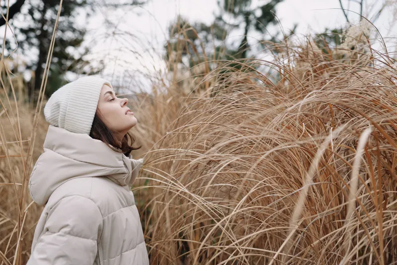 Winter portrait of a woman in a puffer coat and beanie, standing among tall dried grasses and reeds, gazing upward with a calm, thoughtful expression in chilly outdoor scenery.