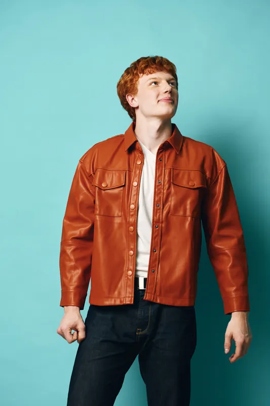 Young man with red hair wearing brown leather jacket and white shirt standing against blue background looking upwards with confident expression in casual style.