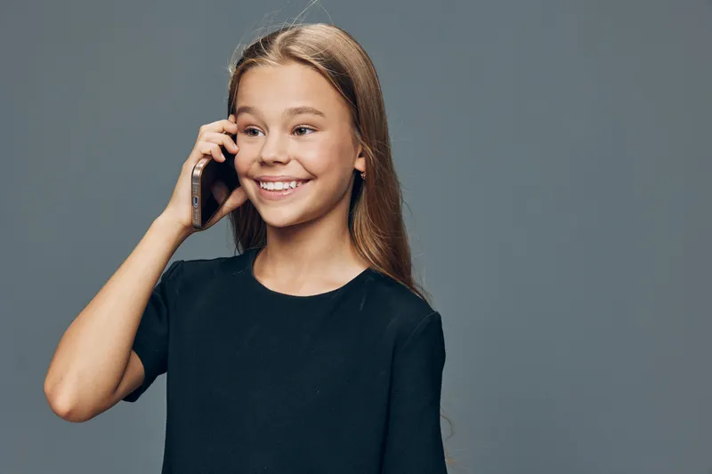 Girl talking on smartphone with smile standing against plain background. Happy child using mobile phone for communication casual lifestyle and technology closeup portrait of female kid.