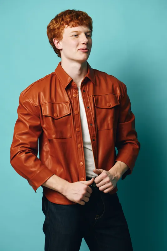 Red-haired man wearing brown leather jacket and white shirt posing in front of blue background. Casual style, confident young male with curly hair and modern outfit.