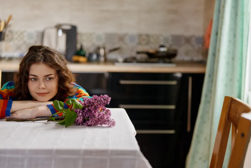 Woman with curly hair resting at a kitchen table, bouquet of flowers nearby, soft daylight on a cozy home setting. A lifestyle moment focused on comfort and wellbeing.