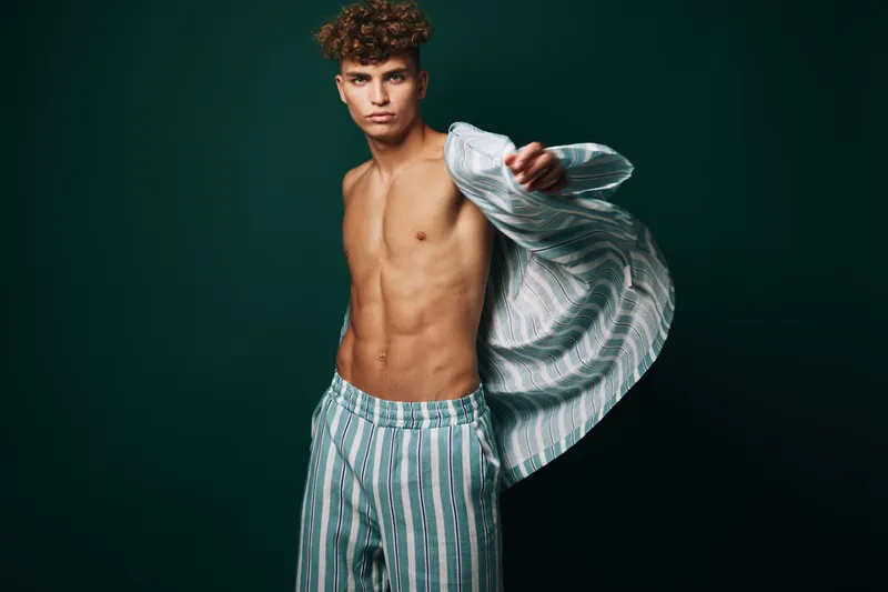 Young man with curly hair wearing striped pajamas shirt open showing toned chest and abs on dark background for fashion and lifestyle concept.