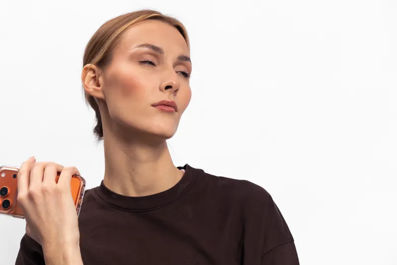 Young woman in brown sweater holding smartphone looks away with closed eyes, expressing calm or contemplation. Studio portrait on white background with soft lighting.
