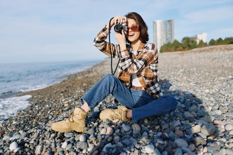 Casual woman with a camera on a pebble beach outdoors, capturing sea and cityscape with a relaxed lifestyle, blending photography, landscape and seaside light