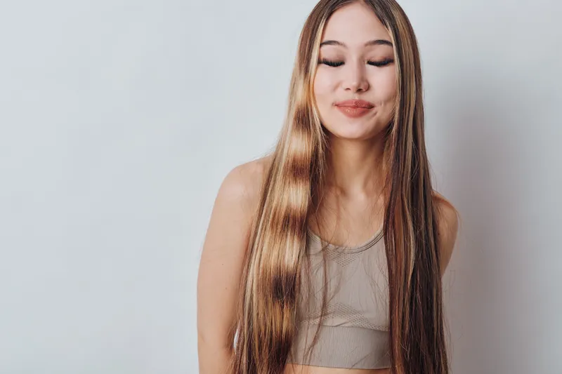Young woman with long straight hair wearing sleeveless top standing on light background with closed eyes and subtle smile for beauty and fashion portrait concept.