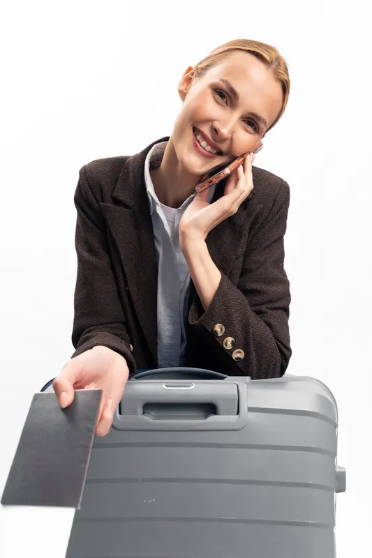 Smiling young woman holding passport and talking on smartphone while leaning on suitcase. Concept of travel, communication, vacation, and happy traveler with modern luggage on white background.