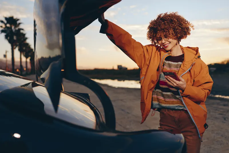 Woman lifestyle car sunset smartphone coastal travel denim jacket smile, curly haired woman by a car at dusk holding a smartphone, orange jacket over denim, warm light for travel independence