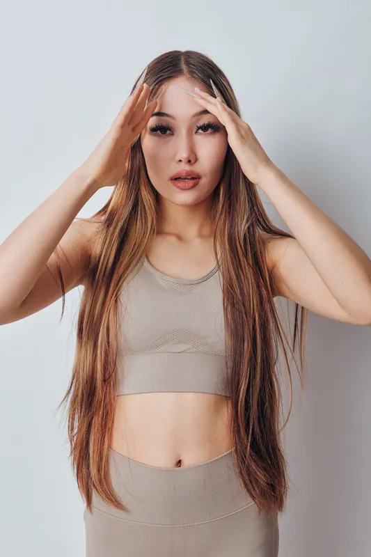 Young woman with long hair in beige top and pants touching forehead with hands, looking stressed or tired, isolated on white background in studio indoors.