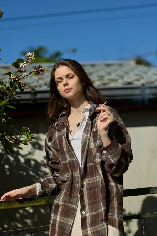 Cheerful woman outdoors, wearing a plaid jacket, leaning on a railing, sunlit moment, relaxed pose, casual streetstyle, natural colors, lush greenery backdrop