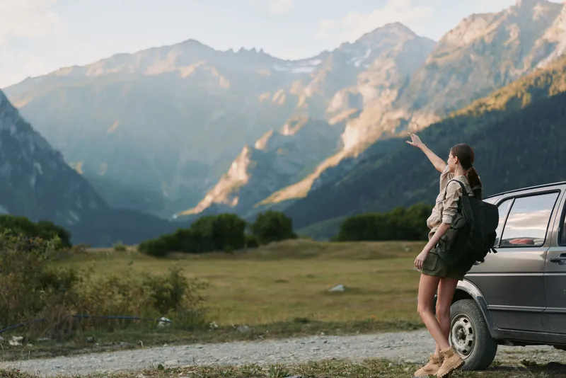 Young woman with a backpack stands beside a car on a mountain road pointing towards the distant peaks during a sunny day in nature.