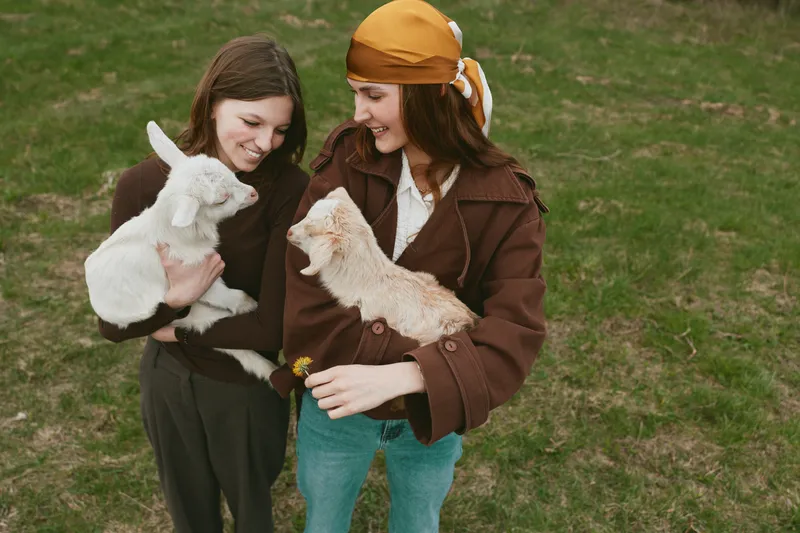 Two women outdoors on a grassy field cuddle with baby goats, showcasing friendship, care, and wholesome lifestyle. Soft daylight, warm smiles, natural setting, simple joy, rustic charm and peaceful