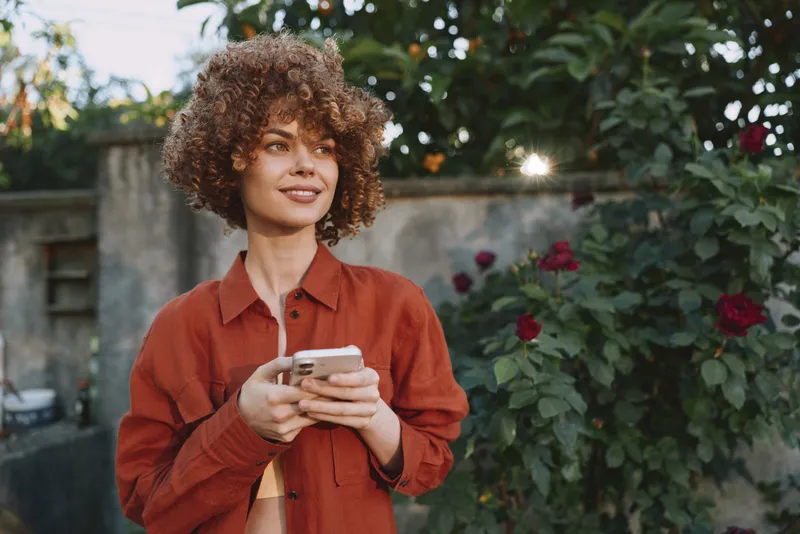 Young woman with curly hair stands outdoors in a red shirt, smiling while holding a smartphone, enjoying casual lifestyle moments in a blooming garden today