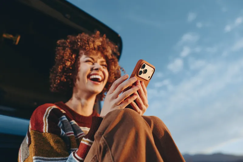 Happy woman lifestyle laughing while taking a selfie with smartphone outdoors near a car, curly hair and cozy sweater, daylight sky, casual travel moment, candid photo, bright mood