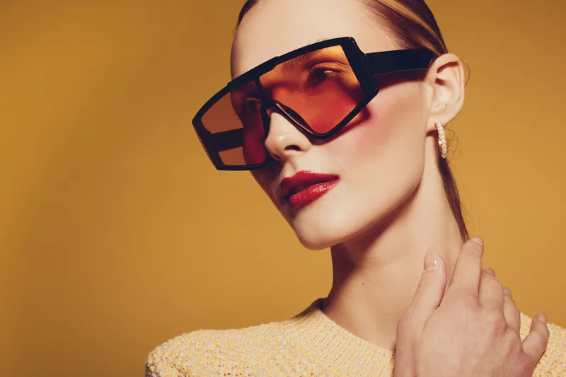 Stylish woman with oversized sunglasses and red lipstick poses against yellow background. Fashion portrait with elegant makeup and trendy accessories. Concept of modern style and beauty.