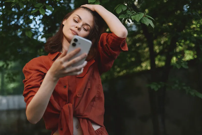 Young woman taking a selfie outdoors, casual fashion, lush greenery in the background, lifestyle moment capturing relaxed confidence and personal style vibe