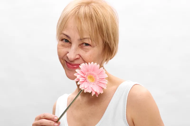 Woman pink flower smile blonde studio white mature portrait cheerful expression, holding a gerbera near her cheek, soft light, clean background, calm mood