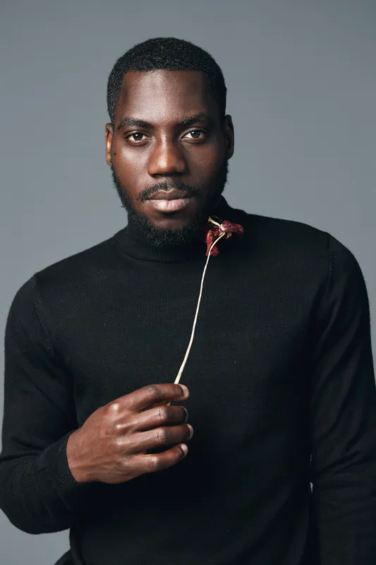 African man with dark skin wearing black turtleneck holding dried flower in creative conceptual visual style studio portrait with serious expression and minimalistic background.