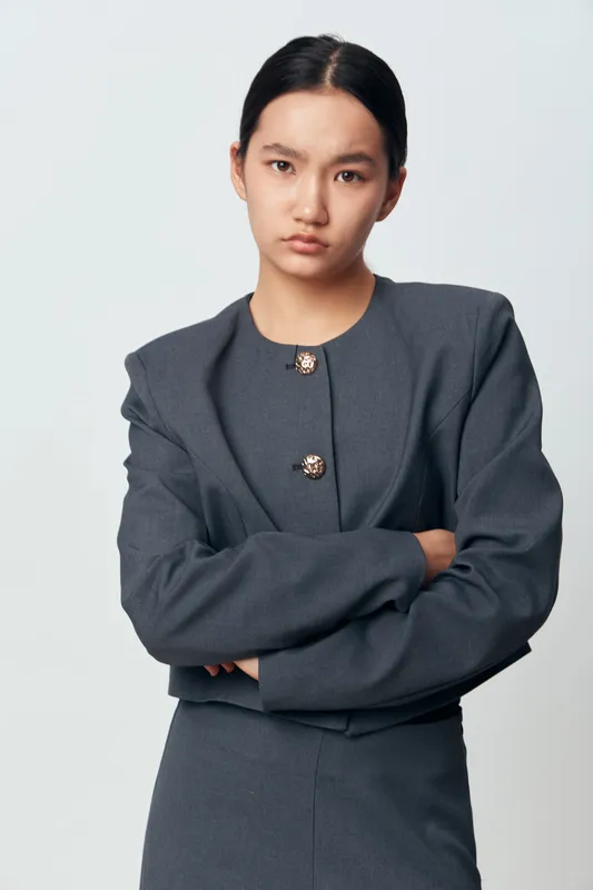 Young woman with crossed arms wearing a formal dark gray suit posing on white background with serious facial expression and straight black hair in professional studio.