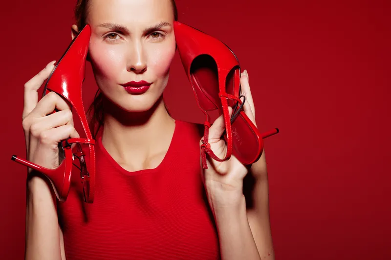 Fashionable woman in red dress holds red high heels near face on red background. Concept of style, elegance, and bold color contrast in fashion photography.