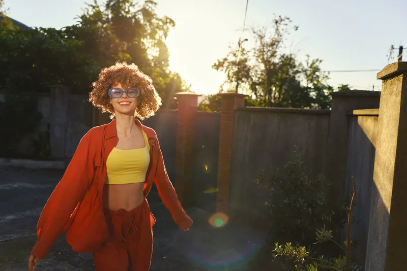 A young woman with curly hair wears a yellow crop top and orange jacket as she walks in a sunlit outdoor space, radiating carefree confidence
