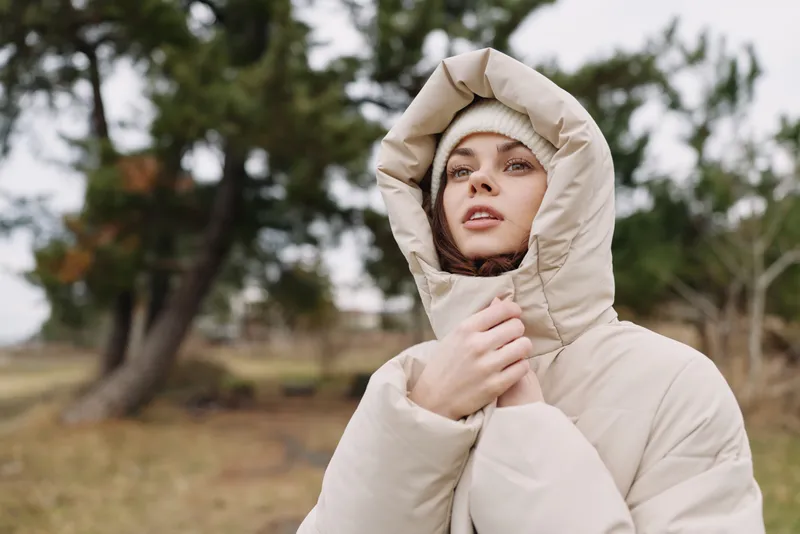 Outdoor portrait of a woman in a bulky hooded coat, looking into the distance with a calm expression in a park during winter light.
