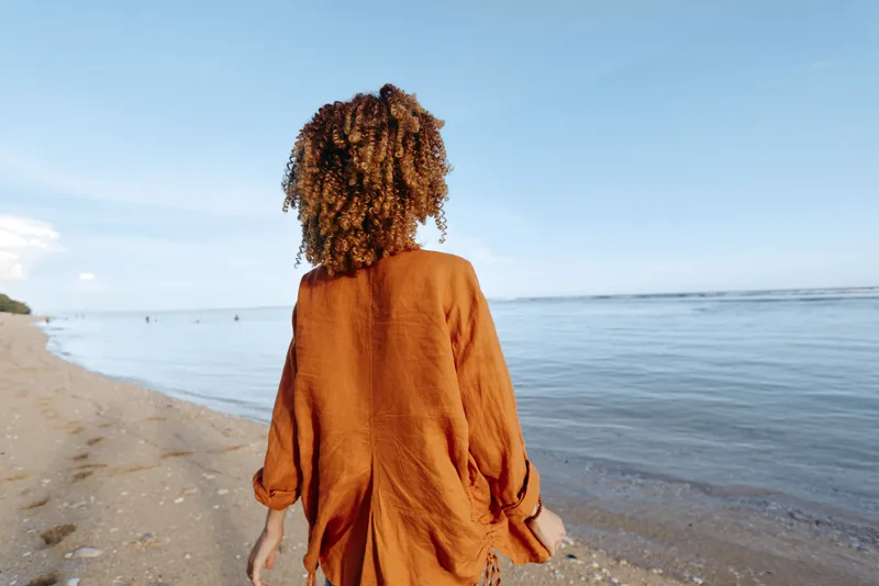 A woman in an orange dress walks along a sunny beach, toward the horizon over ocean.