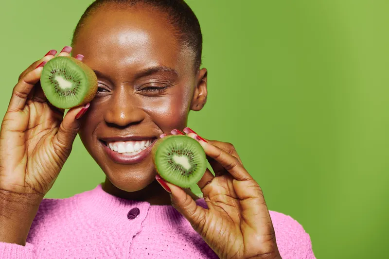Smiling black woman with dark skin holds two halves of kiwi fruit near her face on green background. Concept of healthy lifestyle, fresh food, natural vitamins, and wellness beauty.