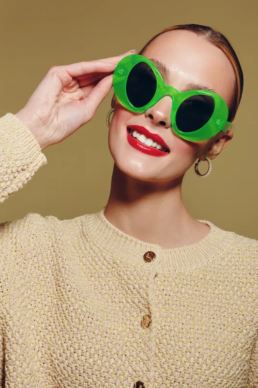 Smiling woman with bright red lipstick wearing large green sunglasses and beige knitted sweater poses against neutral background. Concept of fashion, style, and summer accessories.