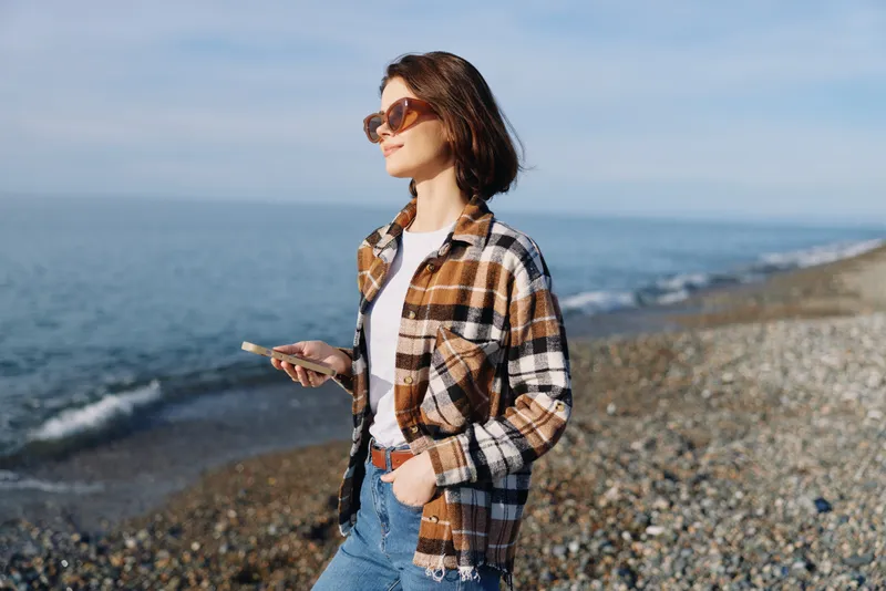 Woman on a pebble beach holding a smartphone, wearing sunglasses and a plaid shirt, casual outdoor style by the sea with a calm horizon and blue sky