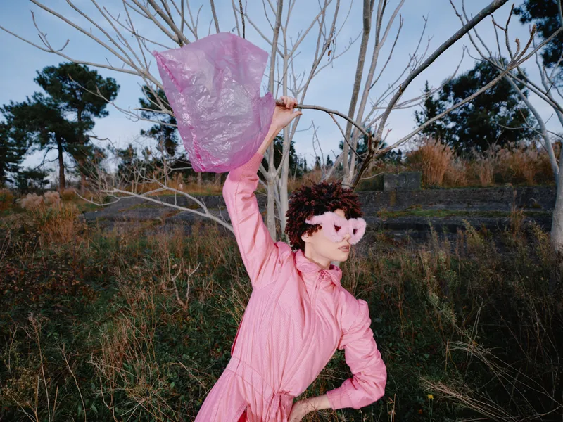 Pink costume performer in an outdoor setting dances and throws a pink scarf, wearing a mask, creating a colorful theatrical moment among bare trees and wind.