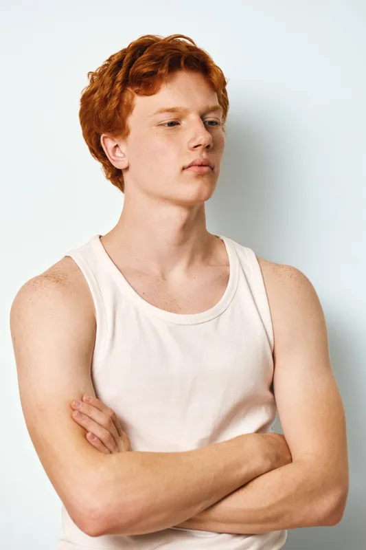 Young man with red hair and freckled skin wearing white tank top looks aside with crossed arms against white background in studio portrait. Casual confident male model.