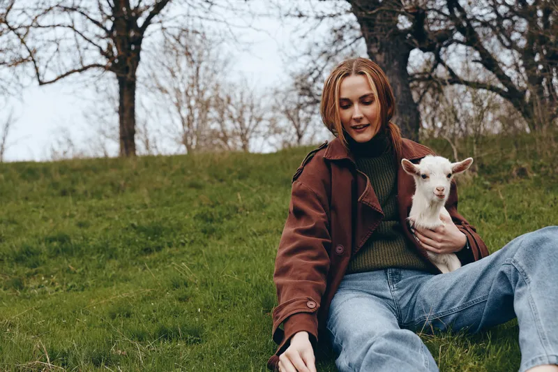 Young woman in a brown jacket sits on green grass holding a white baby goat outdoors in early spring with bare trees in the background, enjoying a calm and natural moment.