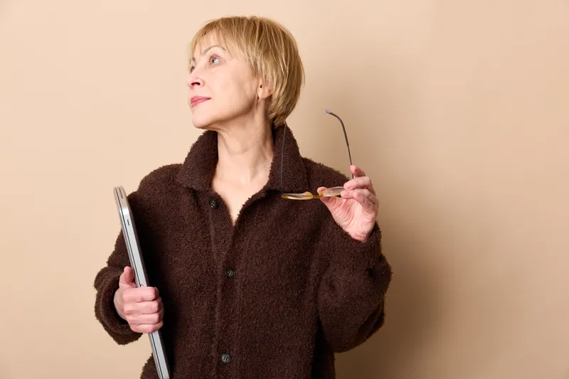 Senior woman studio portrait, holding eyeglasses and glasses near face, thoughtful expression, beige background, brown coat, indoors vision correction for health daily
