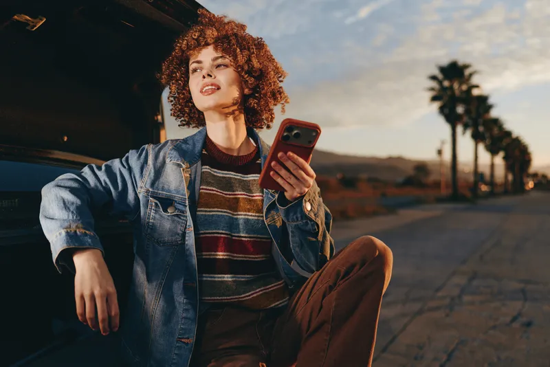 Woman with curly hair wearing denim jacket, sitting outdoors on road, looking at smartphone, casual lifestyle in sunset light, palm trees in background, warm relaxed mood evening