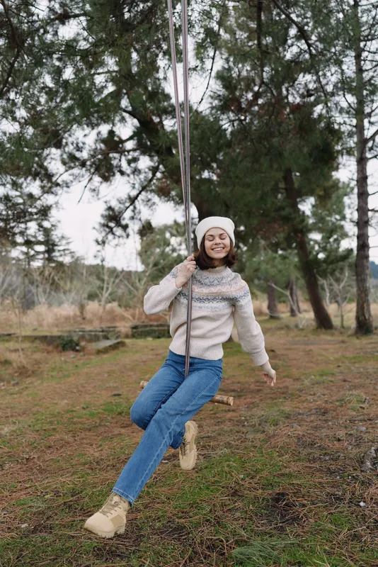 Girl on a swing in a park, enjoying outdoors. Cozy sweater and beanie, smiling as she swings from rope among trees, capturing a carefree childhood moment of joy and play.