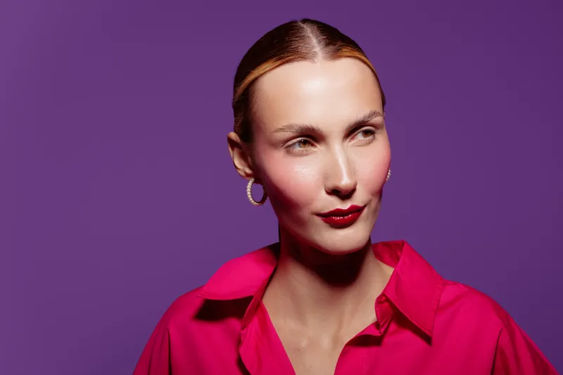 Portrait of confident woman with red lipstick and hoop earrings wearing bright pink shirt against purple background. Concept of fashion, beauty, and modern style with studio lighting.